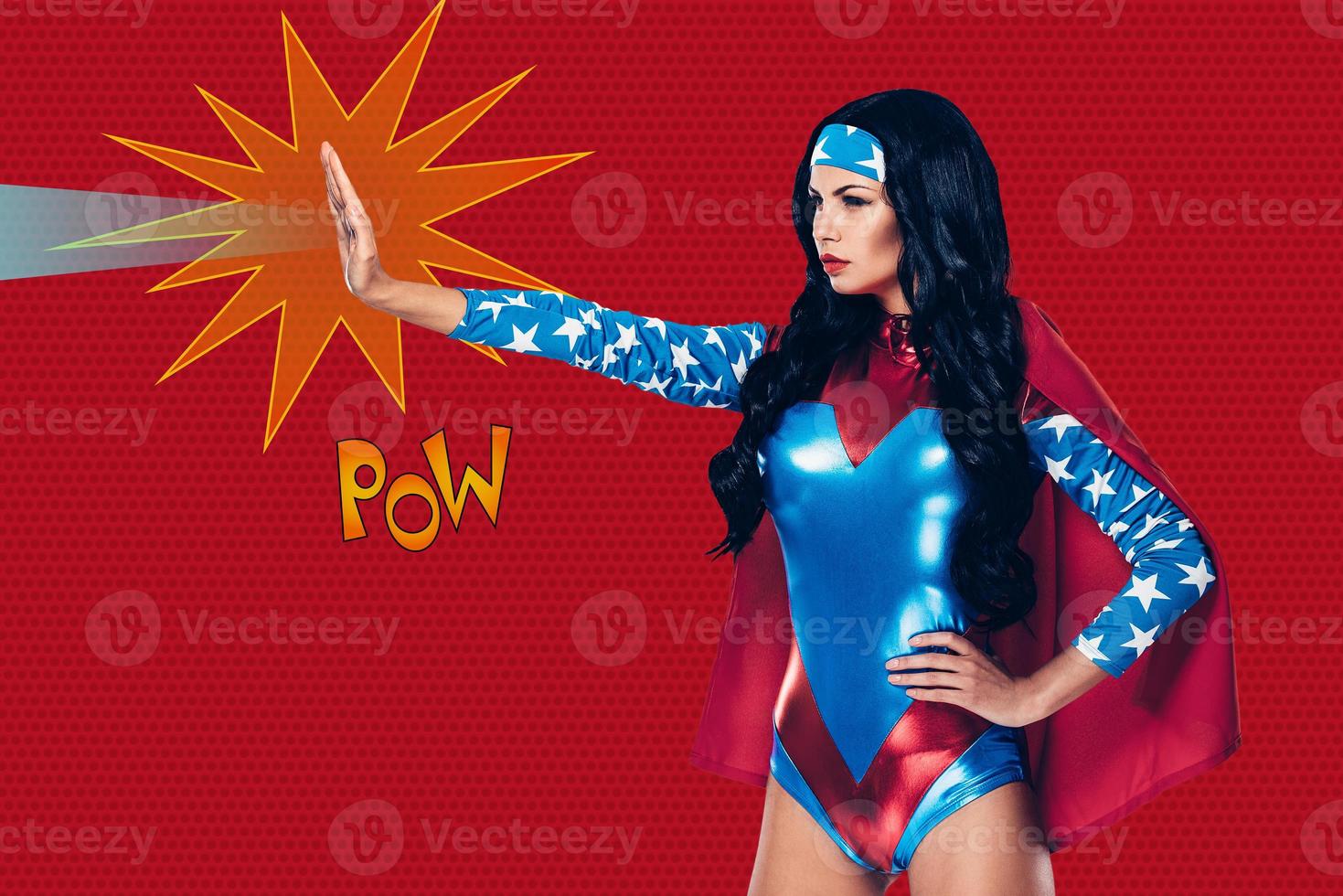 She can stop any crime. Side view of beautiful young woman in superhero costume shooting a beam from her hand while standing against red background photo