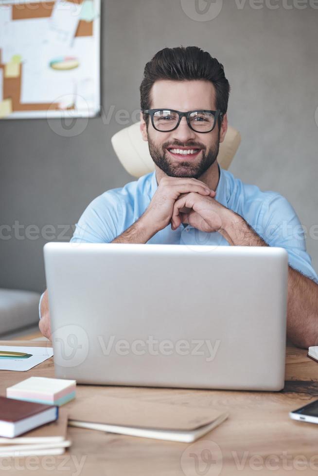 gran solucion alegre joven apuesto hombre con gafas mirando a la cámara con una sonrisa mientras se sienta en su lugar de trabajo foto