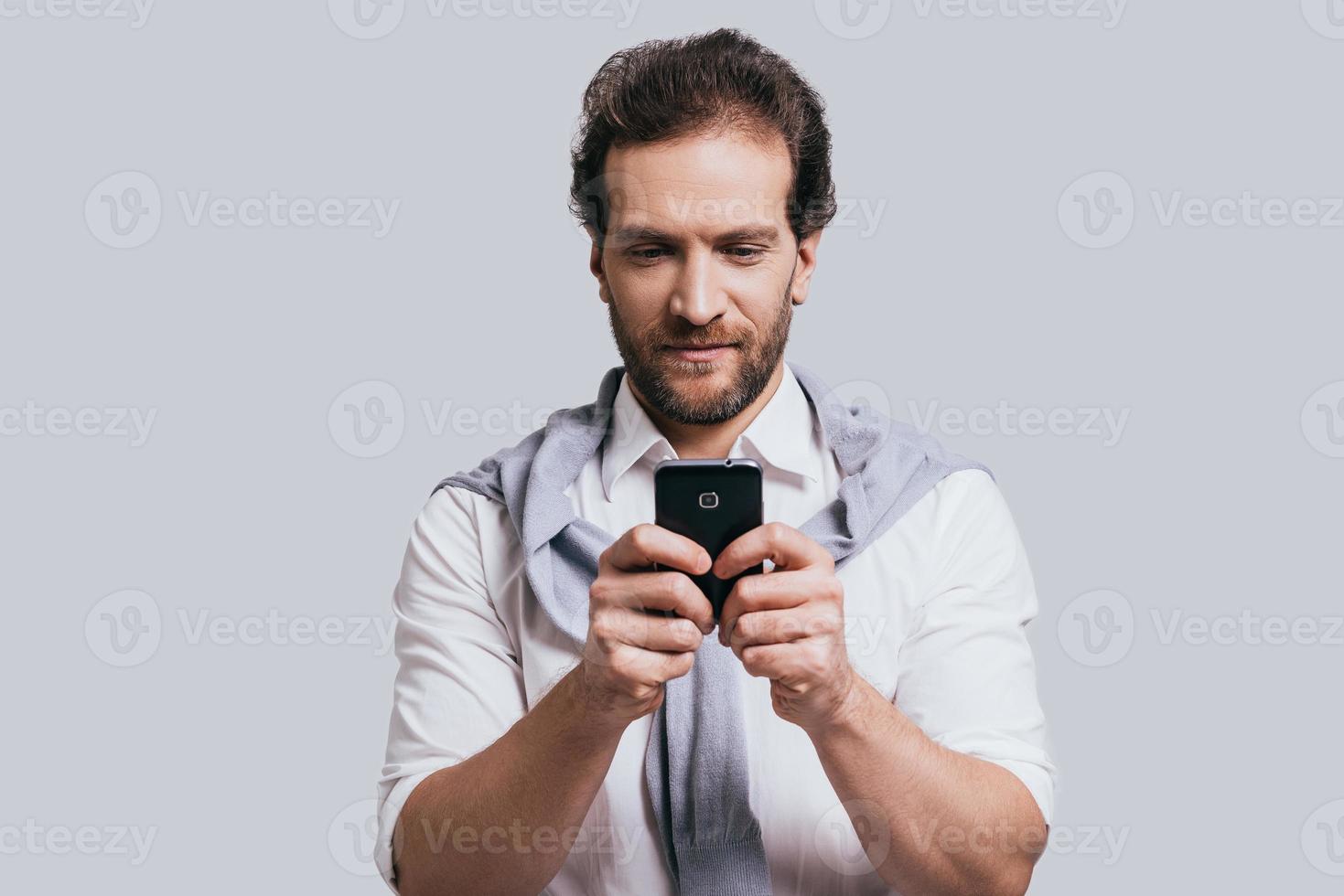 Young and successful. Beautiful young man in smart casual clothes typing a message while standing against grey background photo