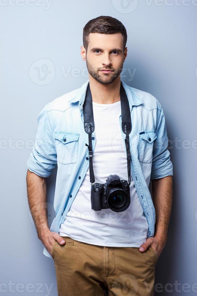 Confident and experienced photographer. Handsome young man with digital camera looking at you while standing against grey background photo