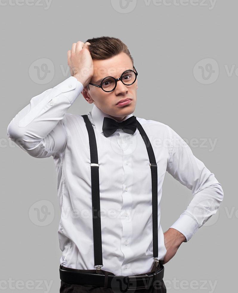 Thoughtful geek. Portrait of thoughtful young man in bow tie and suspenders holding hand in hair and looking away while standing against grey background photo