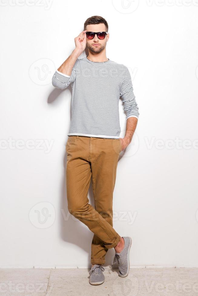 Confident and handsome. Full length of handsome young man adjusting his sunglasses while leaning at the white wall photo