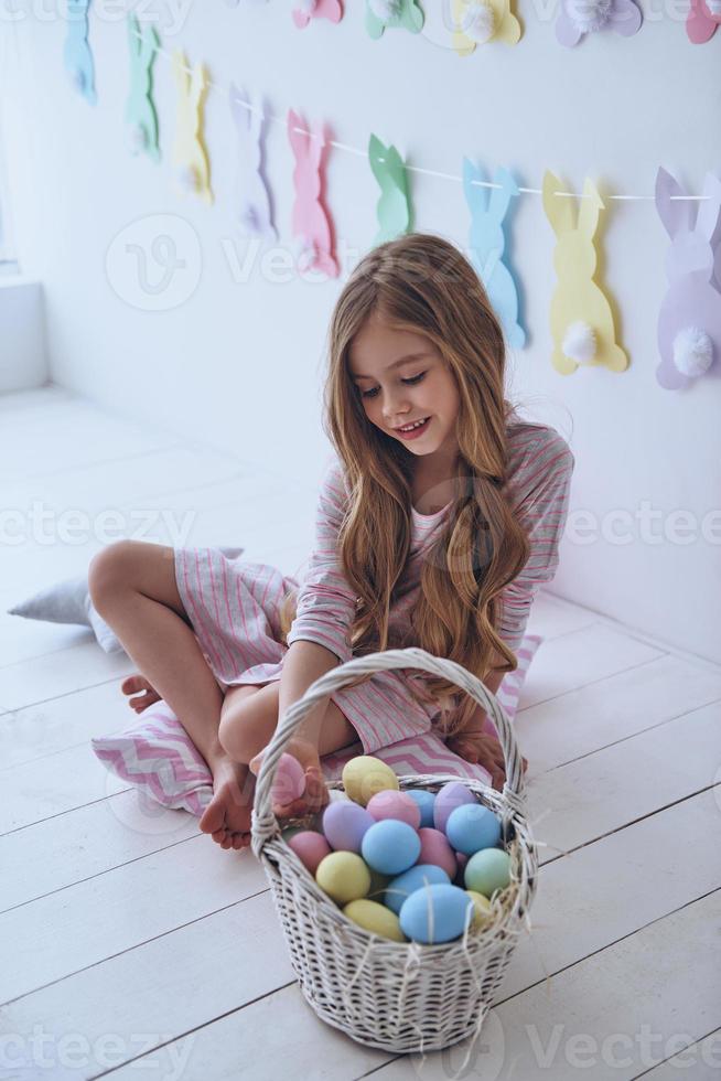 Getting ready to Easter. Lovely little girl holding an Easter egg and smiling while sitting on the pillow with decoration in the background photo