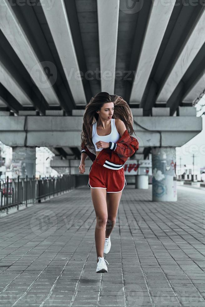 Enjoying her run. Full length of attractive young woman in sport clothing looking away while running under the bridge outdoors photo