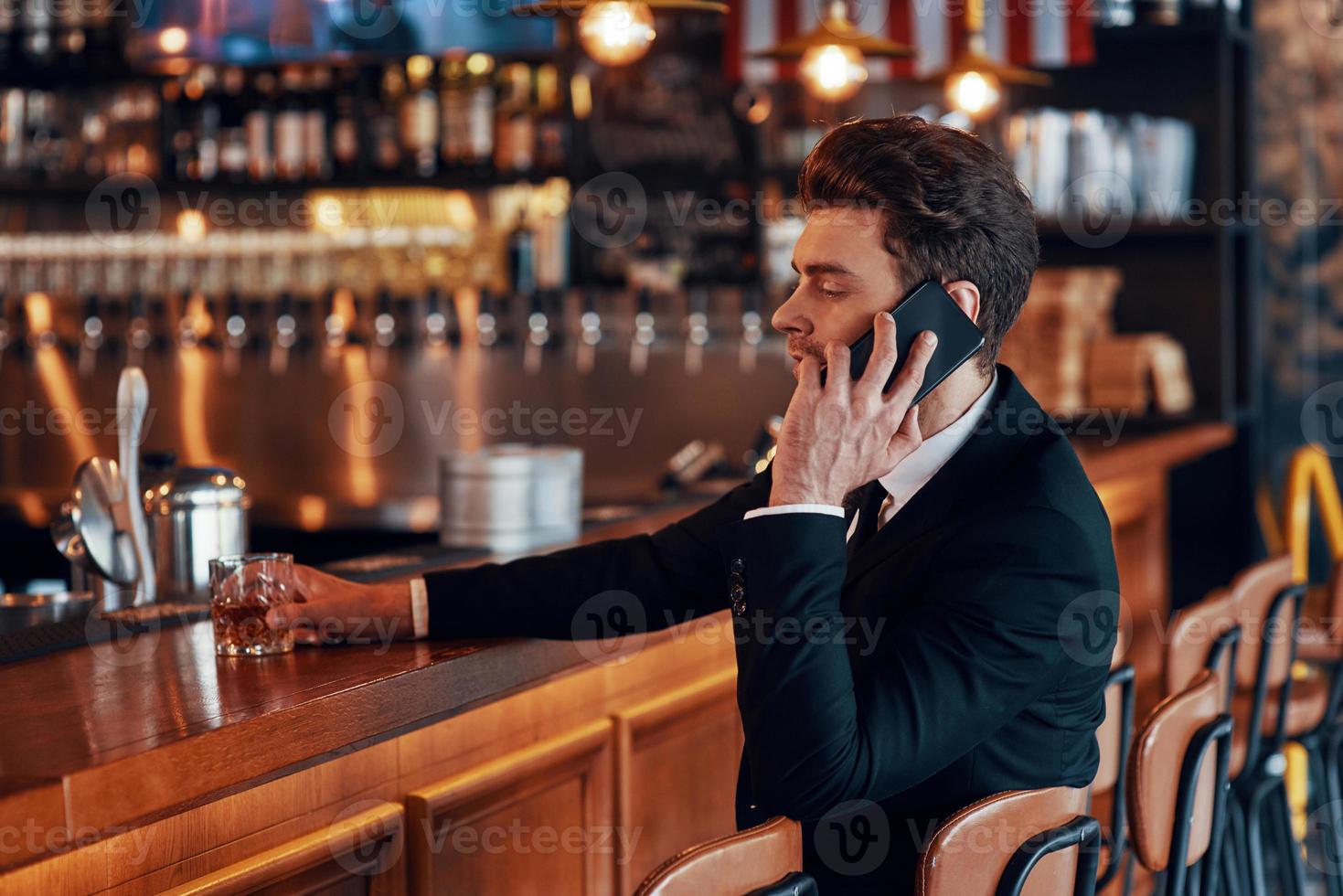 un joven apuesto con traje completo hablando por teléfono inteligente mientras se sienta en la barra del restaurante foto