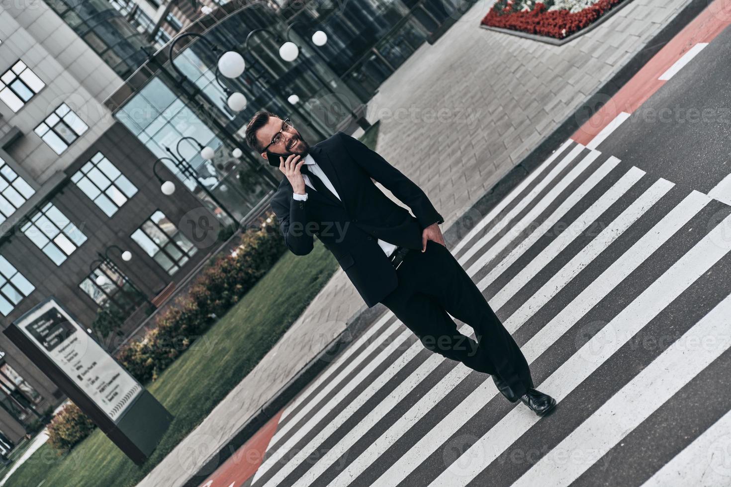 Successful professional. Full length of young man in full suit talking on the phone and smiling while crossing the street photo