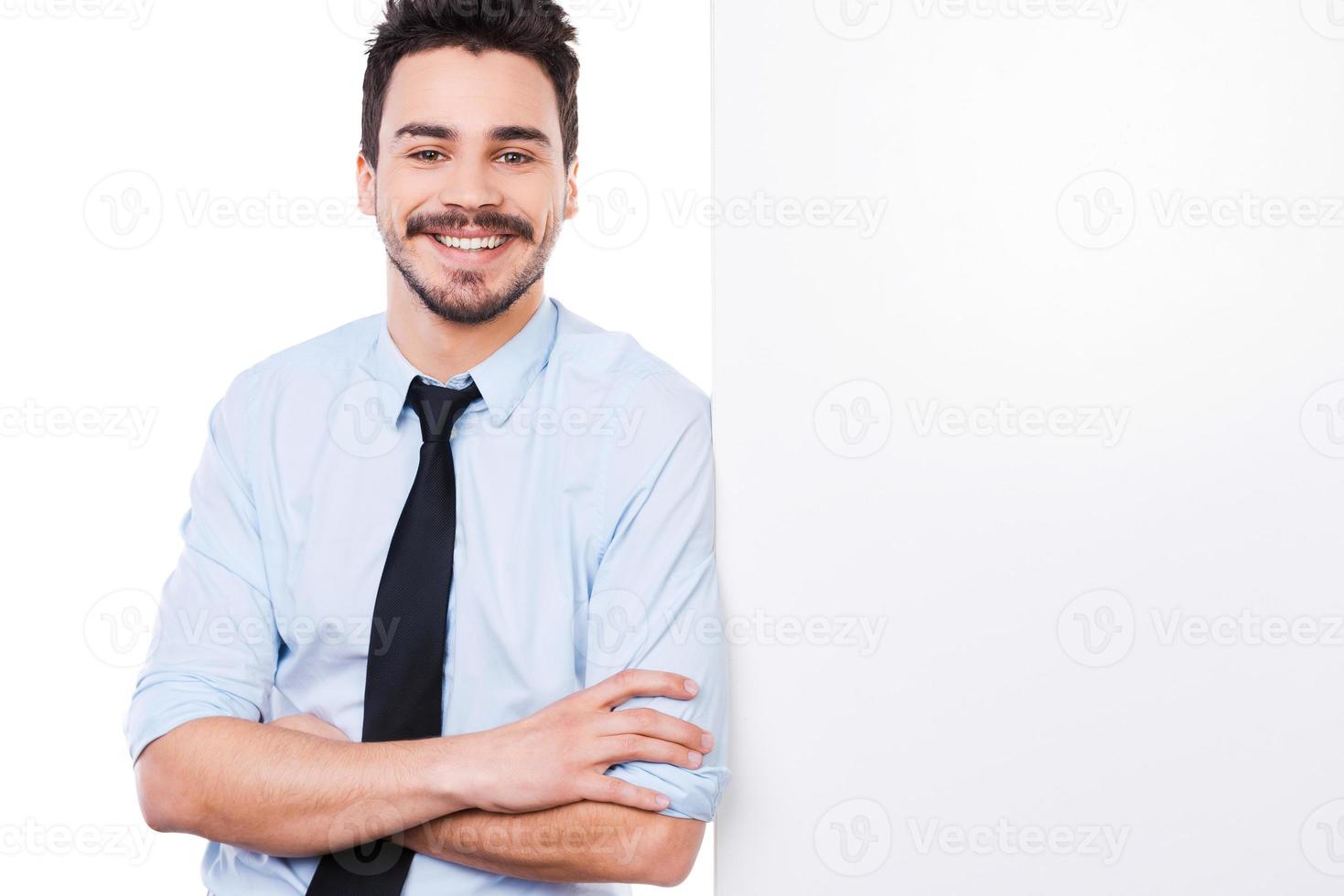 Confident and successful businessman. Handsome young man in shirt and tie leaning at the copy space and keeping arms crossed while standing against white background photo