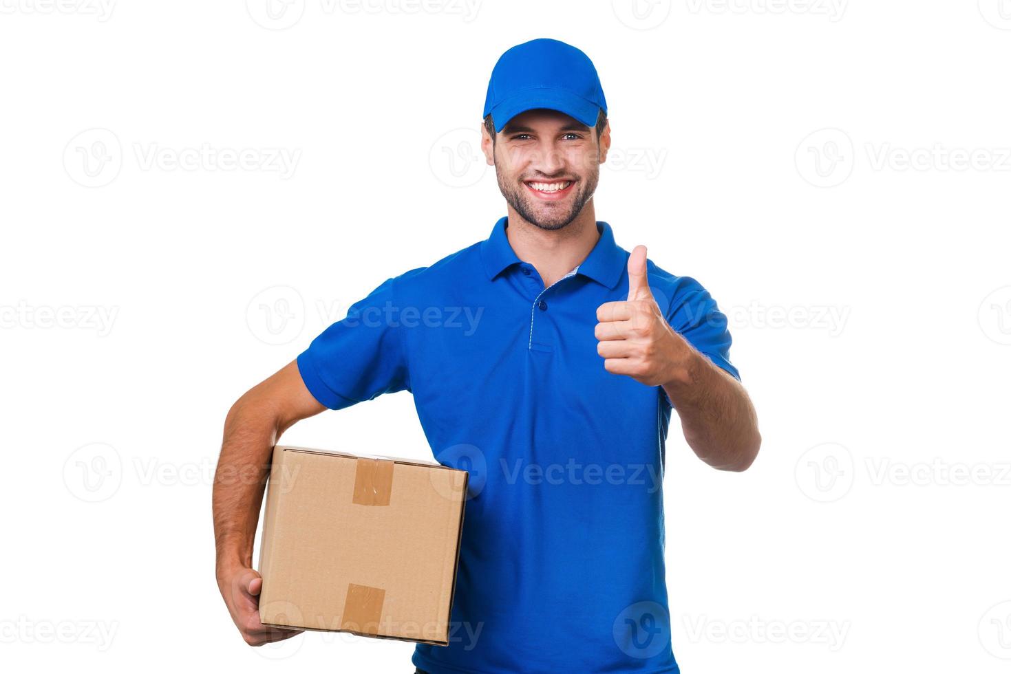 The best delivery service. Cheerful young courier holding a cardboard box and showing his thumb up while standing against white background photo