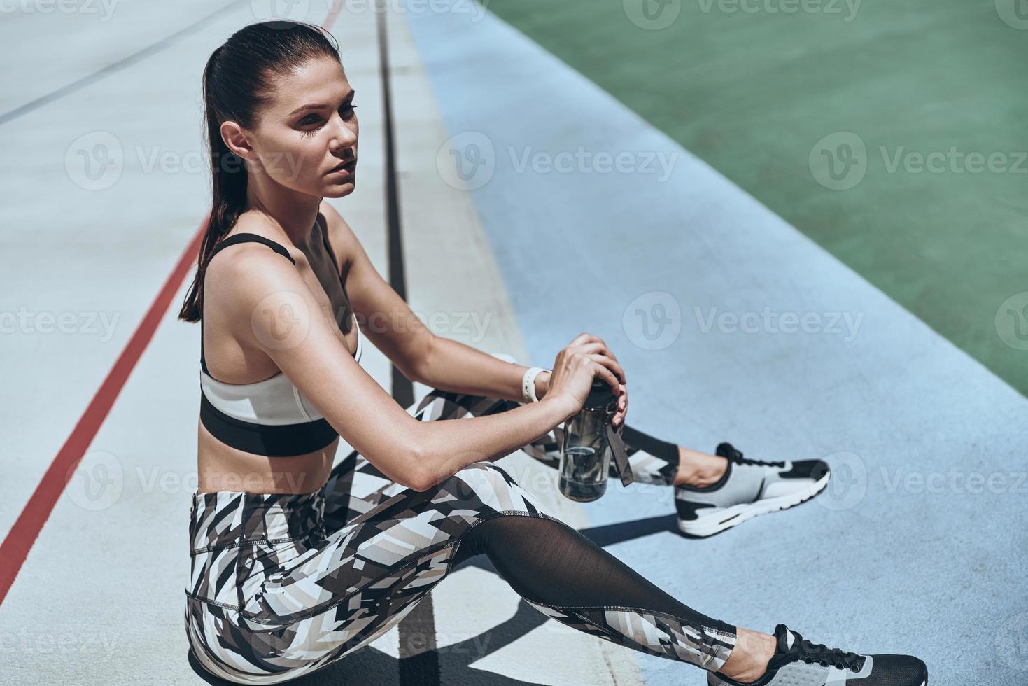 After workout. Top view of beautiful young woman in sports clothing relaxing and looking away while sitting on the running track outdoors photo