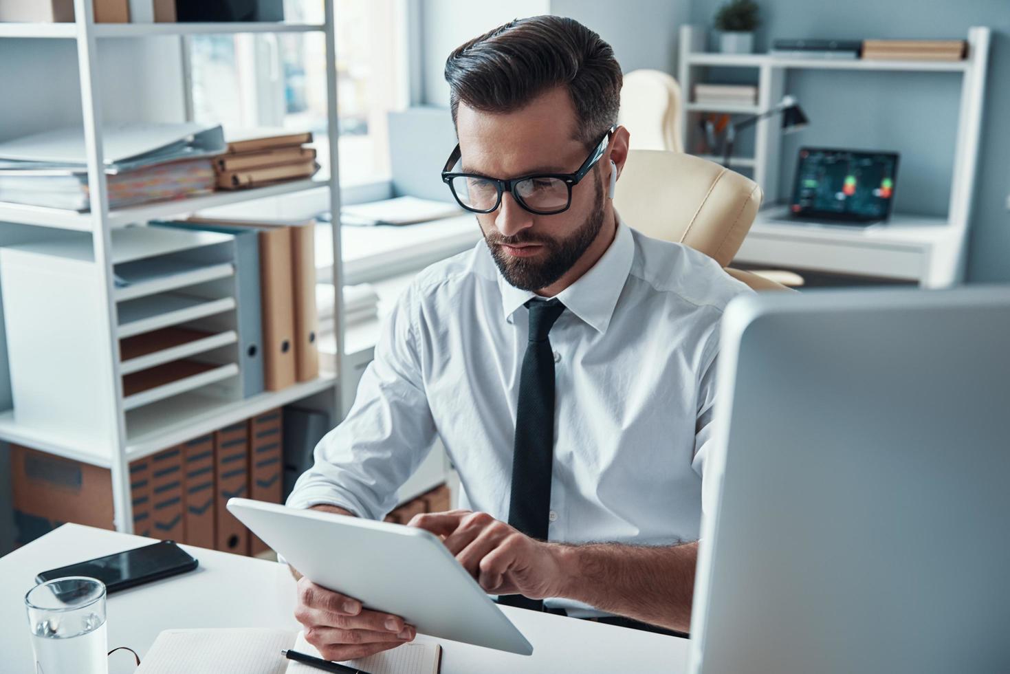 joven ocupado con camisa y corbata trabajando con tecnologías inalámbricas mientras está sentado en la oficina foto