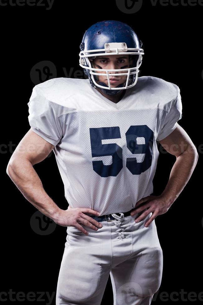 American football player. American football player looking at camera and holding hands on hips while standing against black background photo
