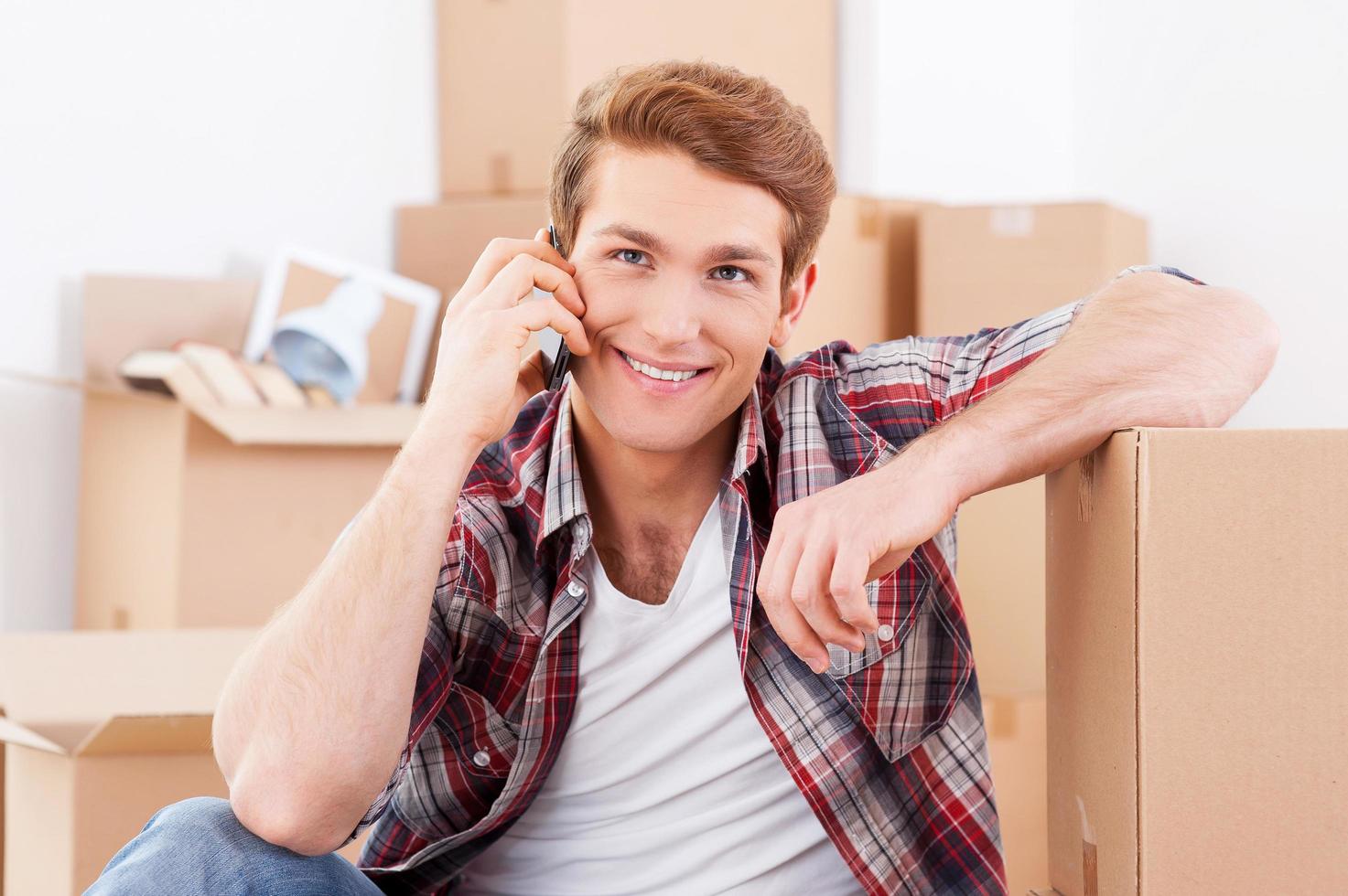 Calling from new house. Handsome young man sitting on the floor and talking on the mobile phone while cardboard boxes laying on the background photo