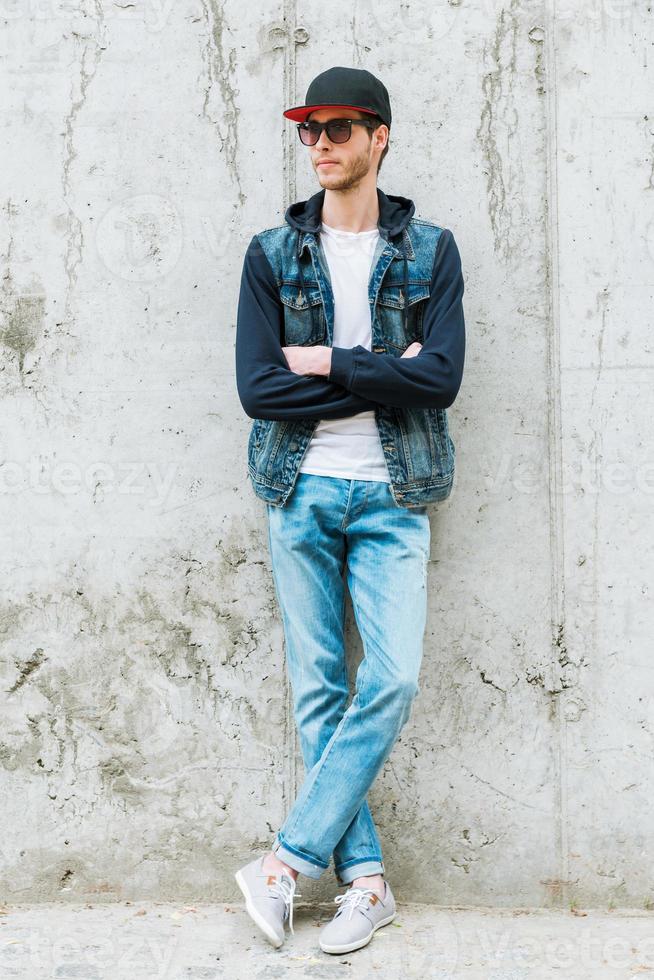Confident style. Handsome young man in headwear keeping arms crossed while standing against concrete wall photo