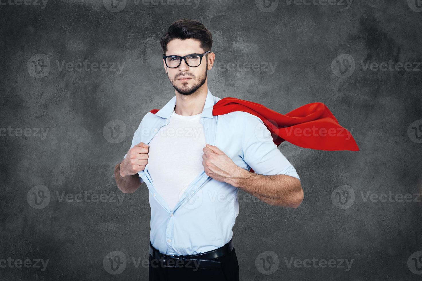 Like a superhero. Confident young man with superhero cape taking of his shirt and looking at camera while stabnding against concrete wall photo