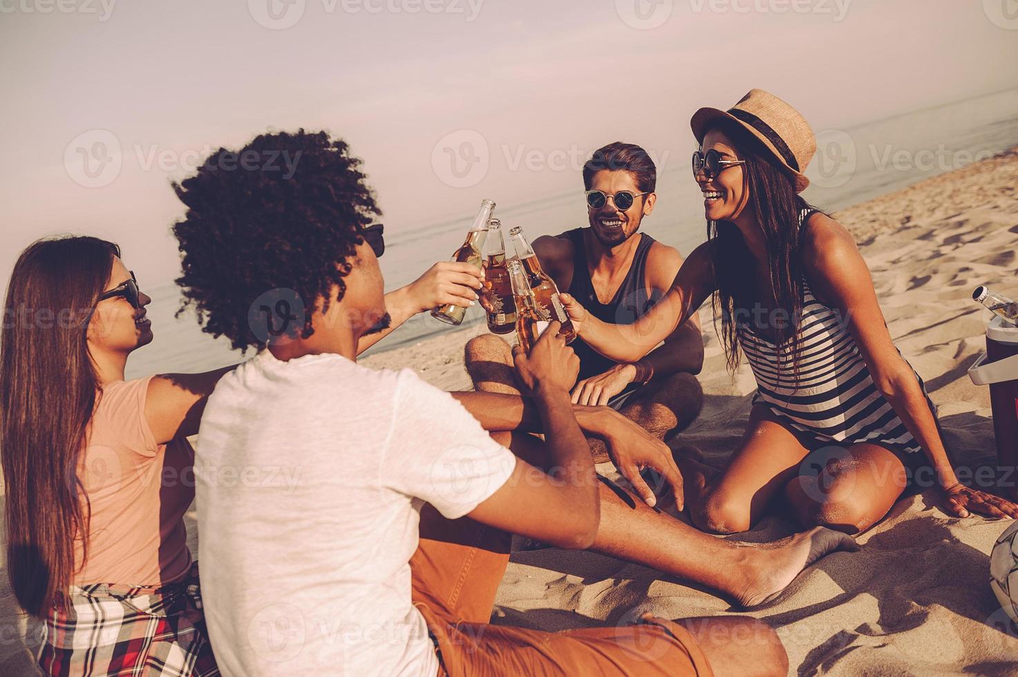 nos saluda a los jóvenes alegres brindando con cerveza y sonriendo mientras nos sentamos juntos en la playa foto