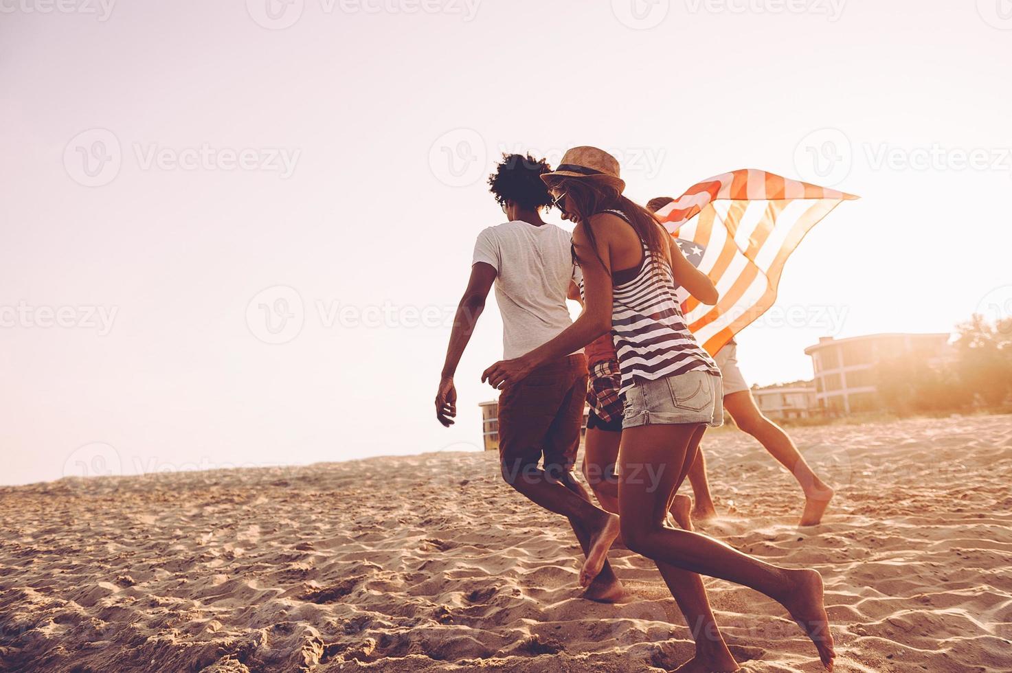 Born to be free. Cheerful young people carrying American flag while running along the beach together photo