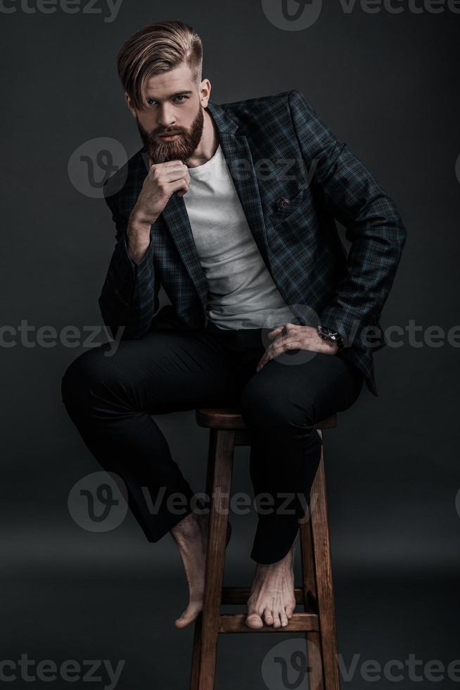 Thoughtful handsome. Full length of stylishly dressed young man looking at camera and holding hand on chin while sitting on chair against grey background photo