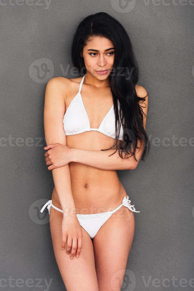 Flirty beauty. Beautiful young mixed race woman in white bikini posing against grey background and looking away photo