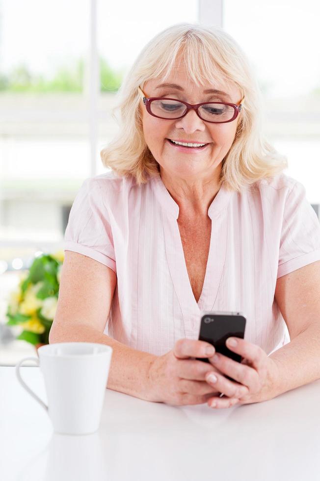 Message from her nearest. Cheerful senior woman looking at her mobile phone and smiling while sitting at the table photo