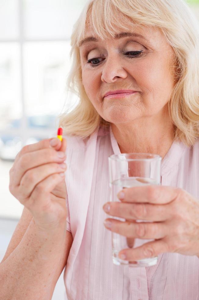 Taking her daily medicines. Depressed senior woman holding pill and glass of water while sitting indoors photo