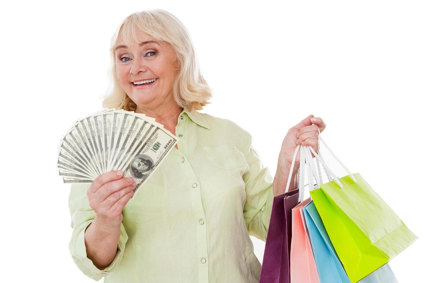 adicto a las compras mayor. feliz anciana estirando la mano con papel moneda y agujereando bolsas de compras mientras se encuentra aislada en el fondo blanco foto