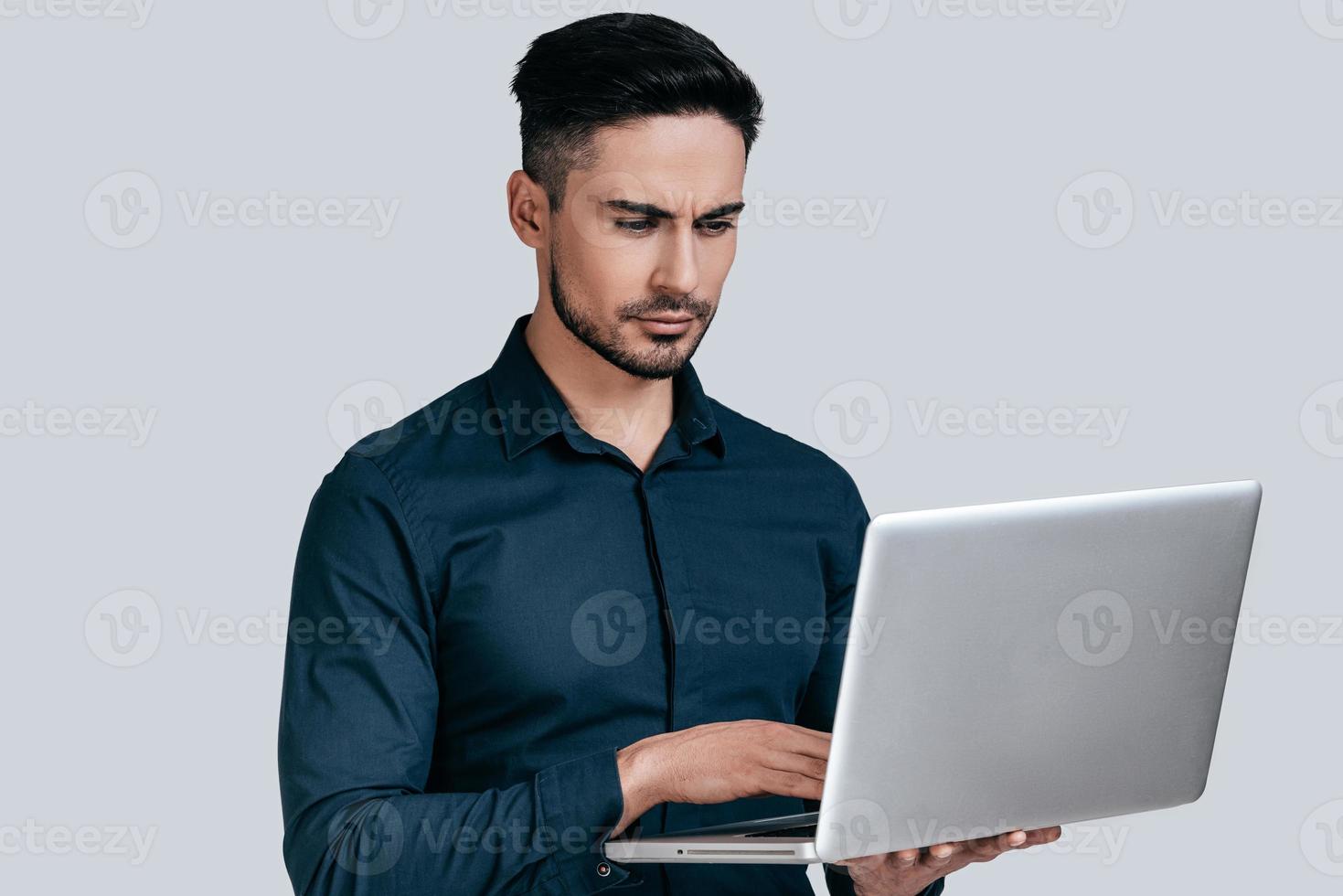 Concentrated at work.  Good looking young man in shirt working on laptop while standing against grey background photo