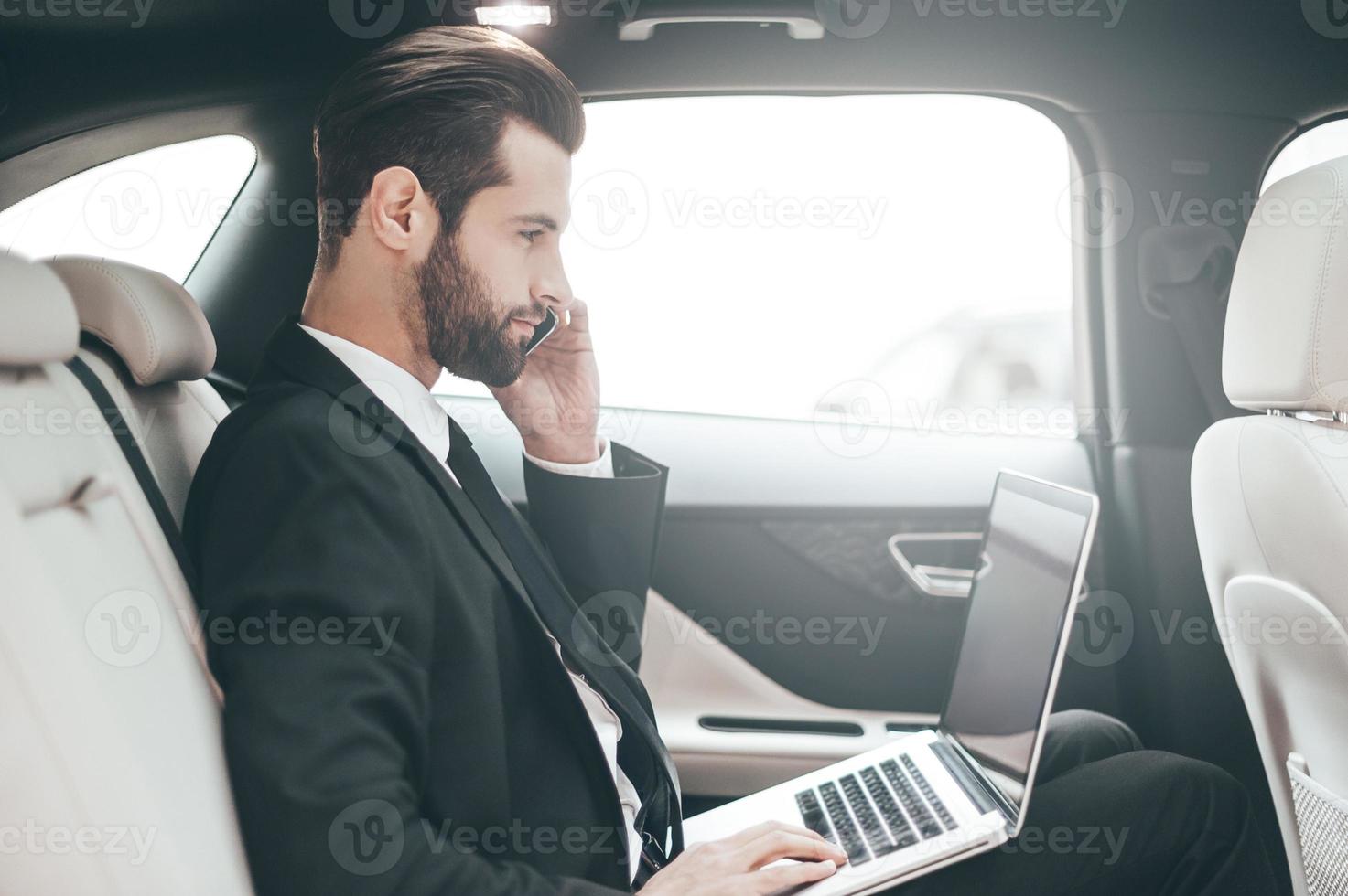 ocupado en el trabajo. joven hombre de negocios confiado trabajando en su laptop y hablando por teléfono mientras está sentado en el auto foto