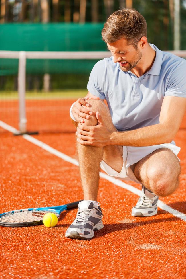 Feeling pain in knee. Close-up of tennis player touching his knee and grimacing while sitting on the tennis court photo