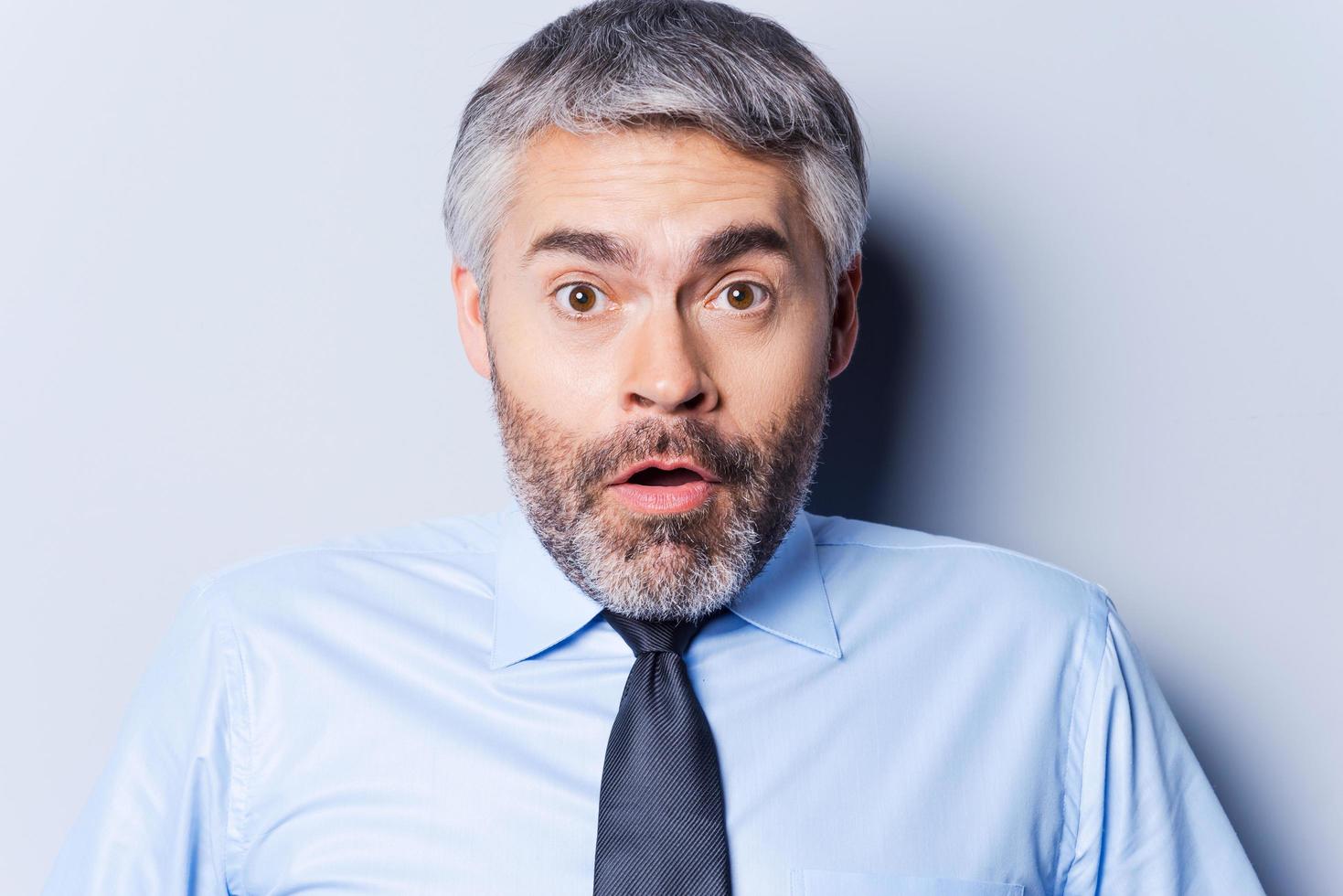 Surprised businessman. Surprised mature man in shirt and tie staring at camera and keeping mouth open while standing against grey background photo