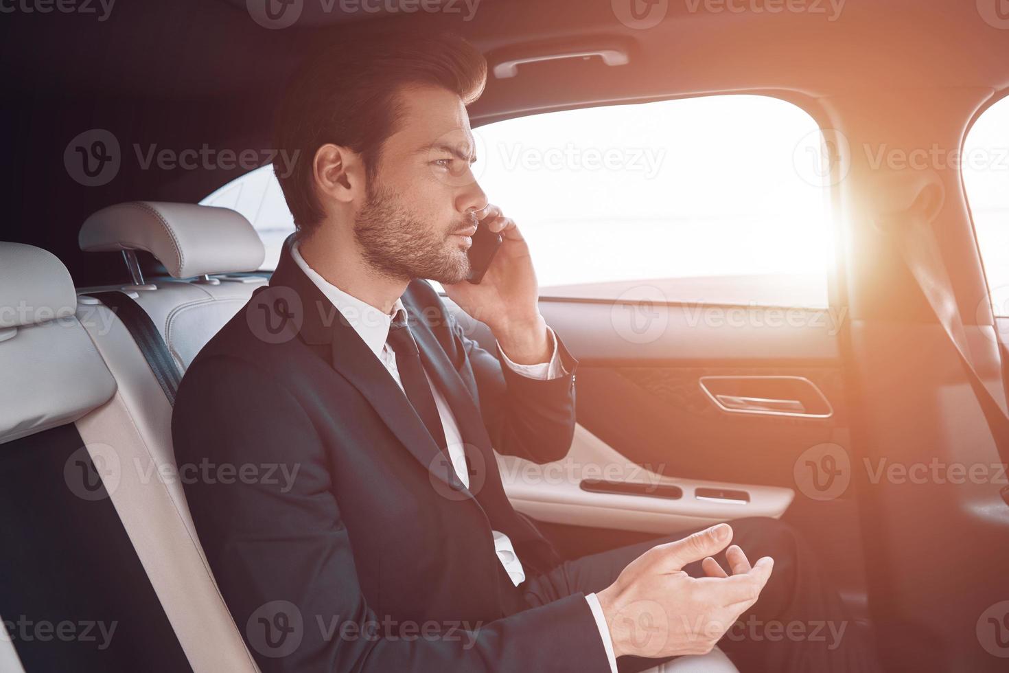 siempre disponible. un joven apuesto con traje completo hablando por teléfono inteligente mientras está sentado en el auto foto