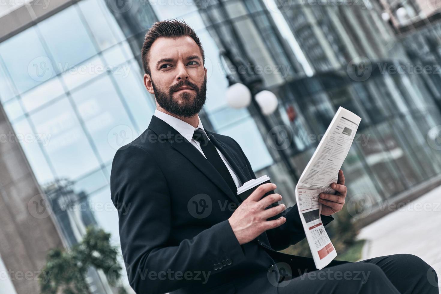 confiado e inteligente. un joven apuesto con traje completo leyendo un periódico mientras se sienta al aire libre foto