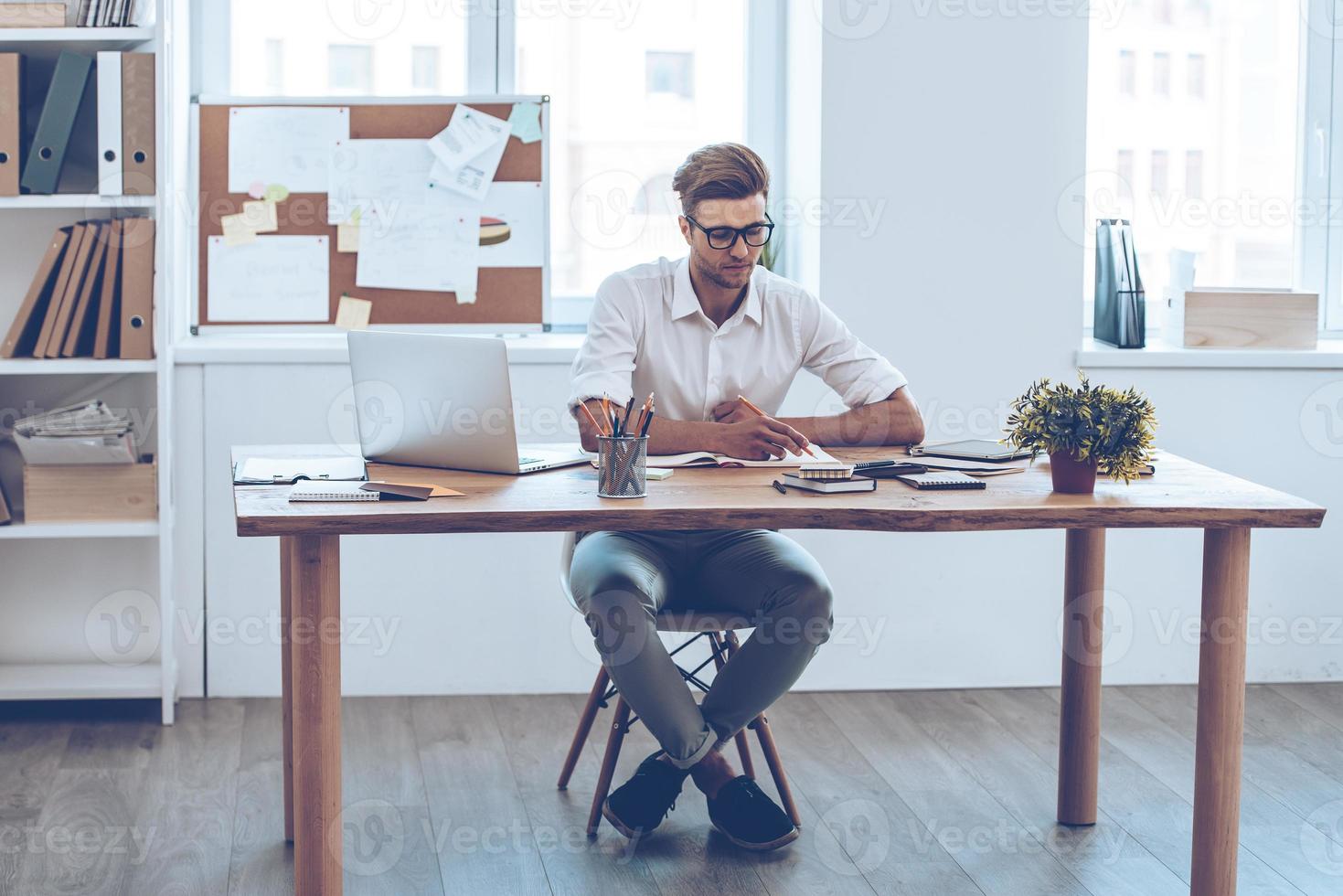 Making some notes. Pensive young handsome man writing in his notebook while sitting at his working place photo