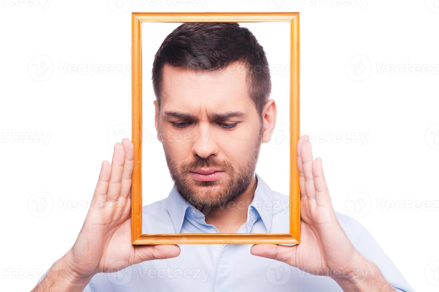 Depressive portrait. Portrait of frustrated young man in shirt holding picture frame in front of his face  while standing against white background photo