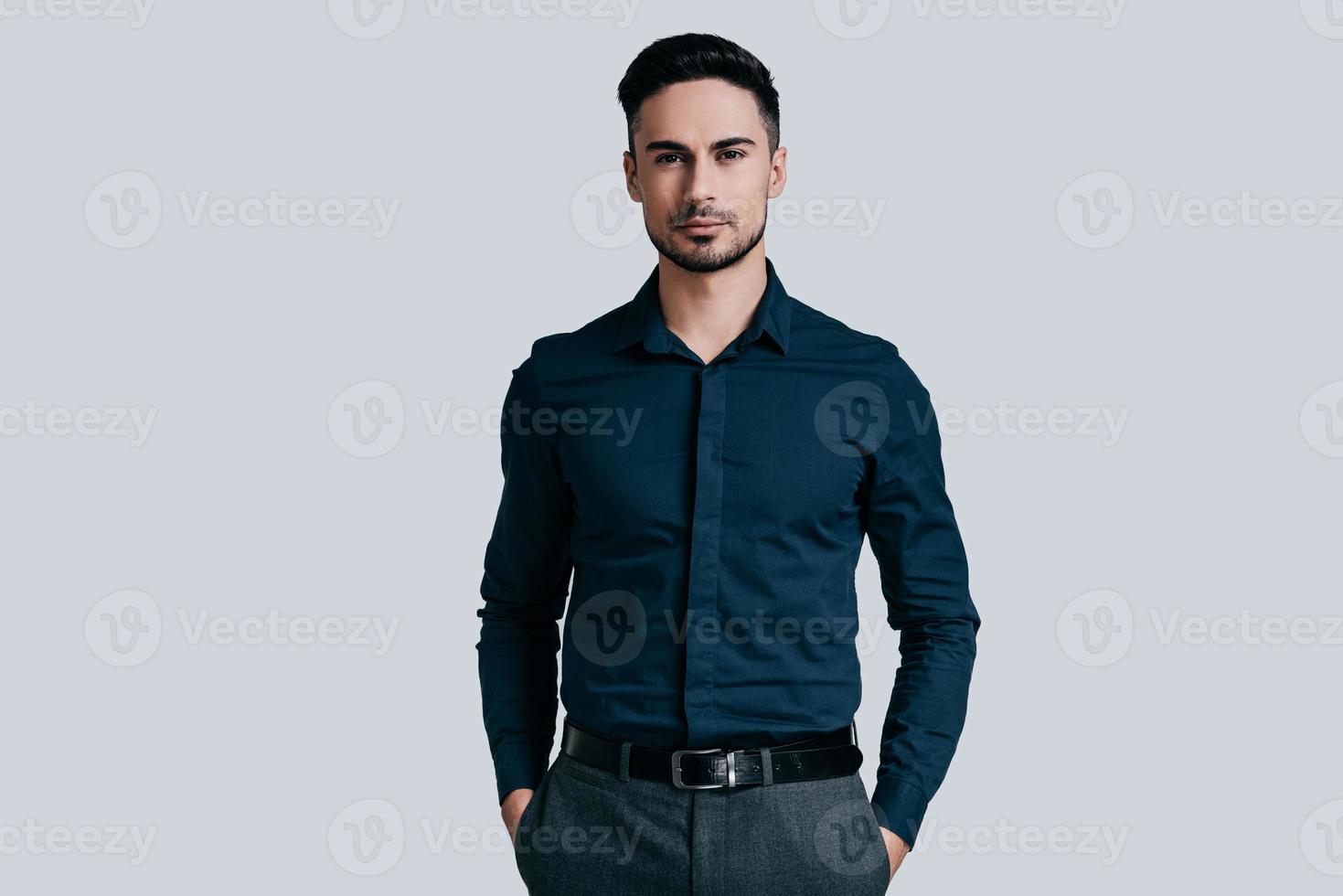 Confidence and charisma. Good looking young man keeping hands in pockets and looking at camera while standing against grey background photo