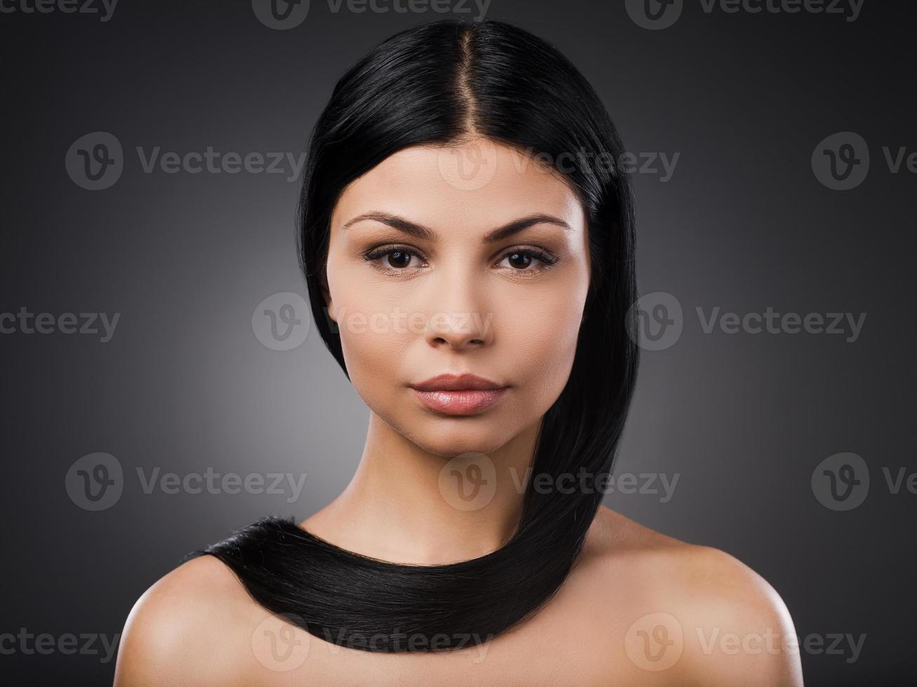 cabello largo y saludable. bella joven sin camisa mirando a la cámara y cubriendo su cuello con el pelo mientras se enfrenta a un fondo gris foto
