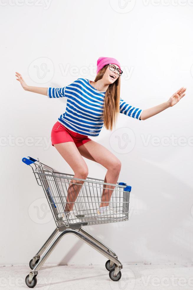 compras extremas. Una joven juguetona con sombreros y gafas montando en un carrito de compras con fondo gris foto