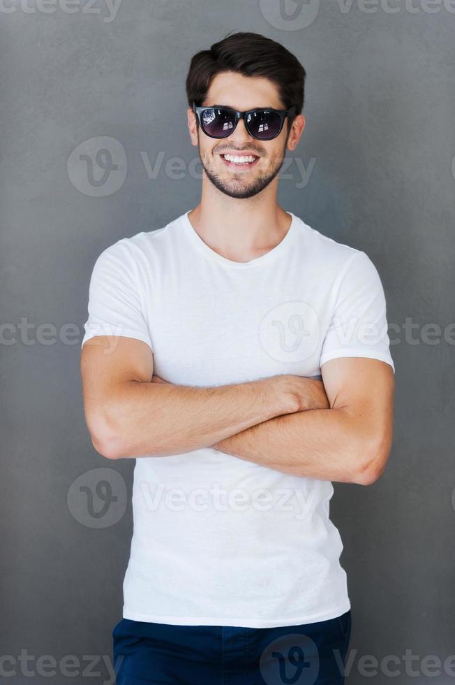 Casually handsome. Handsome young man keeping arms crossed and smiling while standing against grey background photo