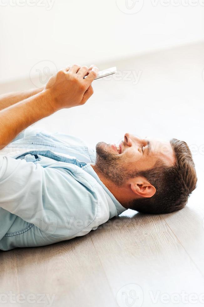 Taking time to relax. Side view of young man holding mobile phone and smiling while lying on the floor at his apartment photo