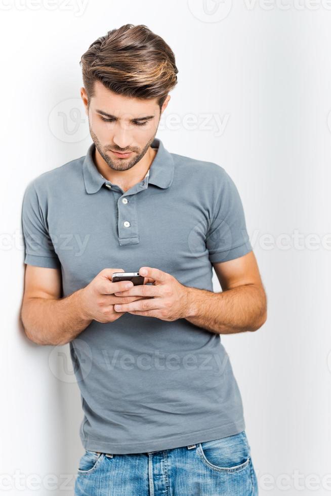 Texting to friend. Handsome young man holding mobile phone and looking at it while leaning at the wall photo