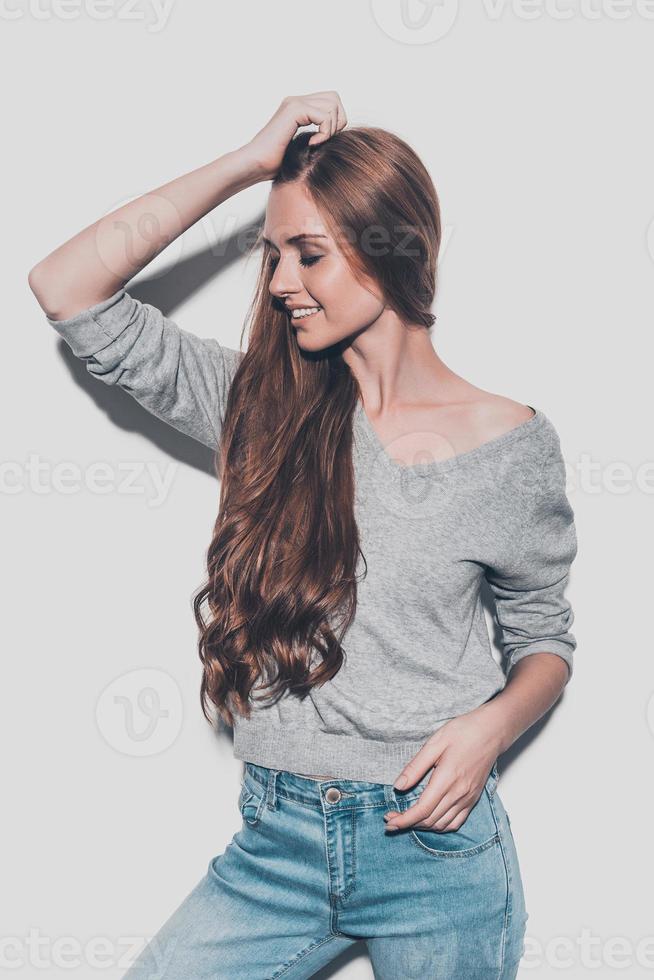 orgullosa de su cabello saludable. Atractiva joven mujer sonriente sosteniendo la mano en el cabello y manteniendo los ojos cerrados mientras se enfrenta a un fondo gris foto