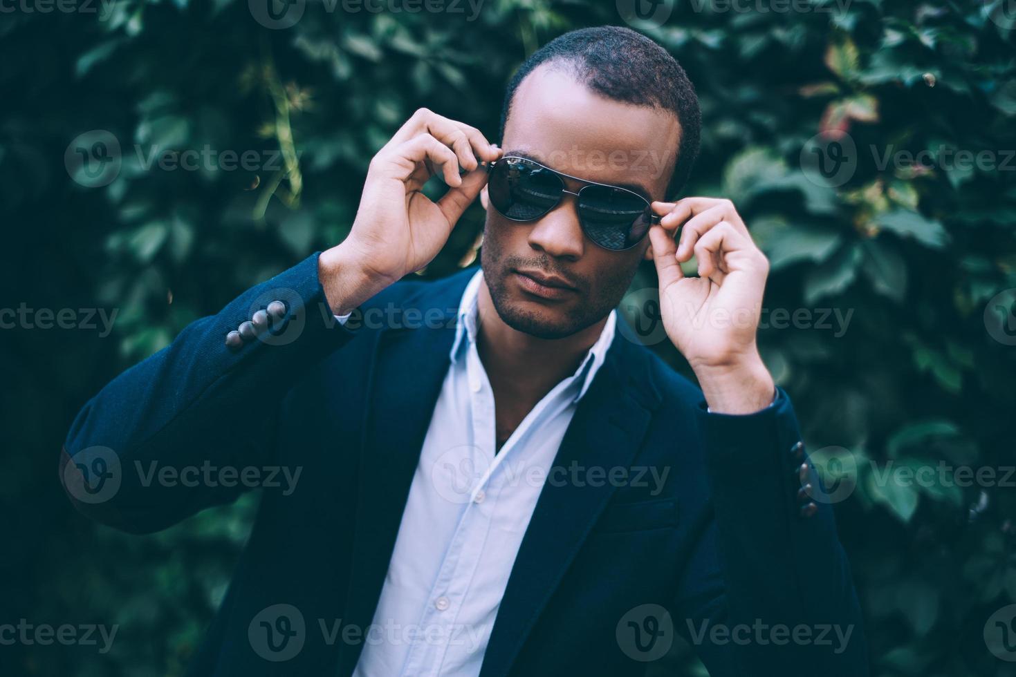 confiado en su estilo perfecto. un apuesto joven africano con ropa informal inteligente ajustando sus gafas de sol y mirando la cámara mientras se enfrenta al fondo de una planta verde al aire libre foto