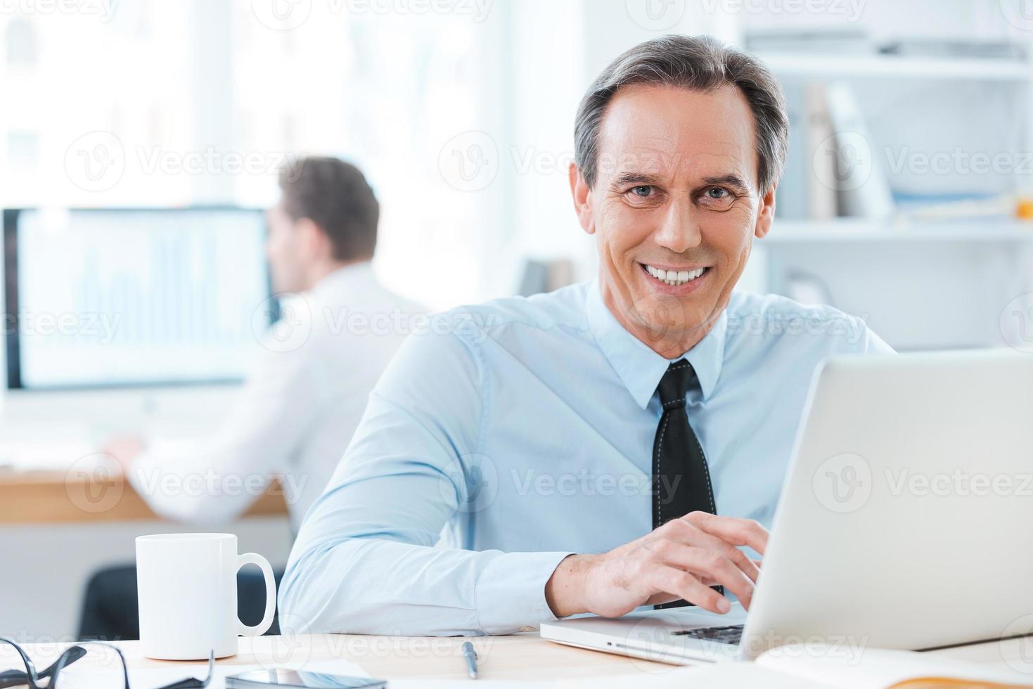 He thrives in this work. Happy businessman in formalwear working on laptop and smiling at camera while sitting at his desk in office photo