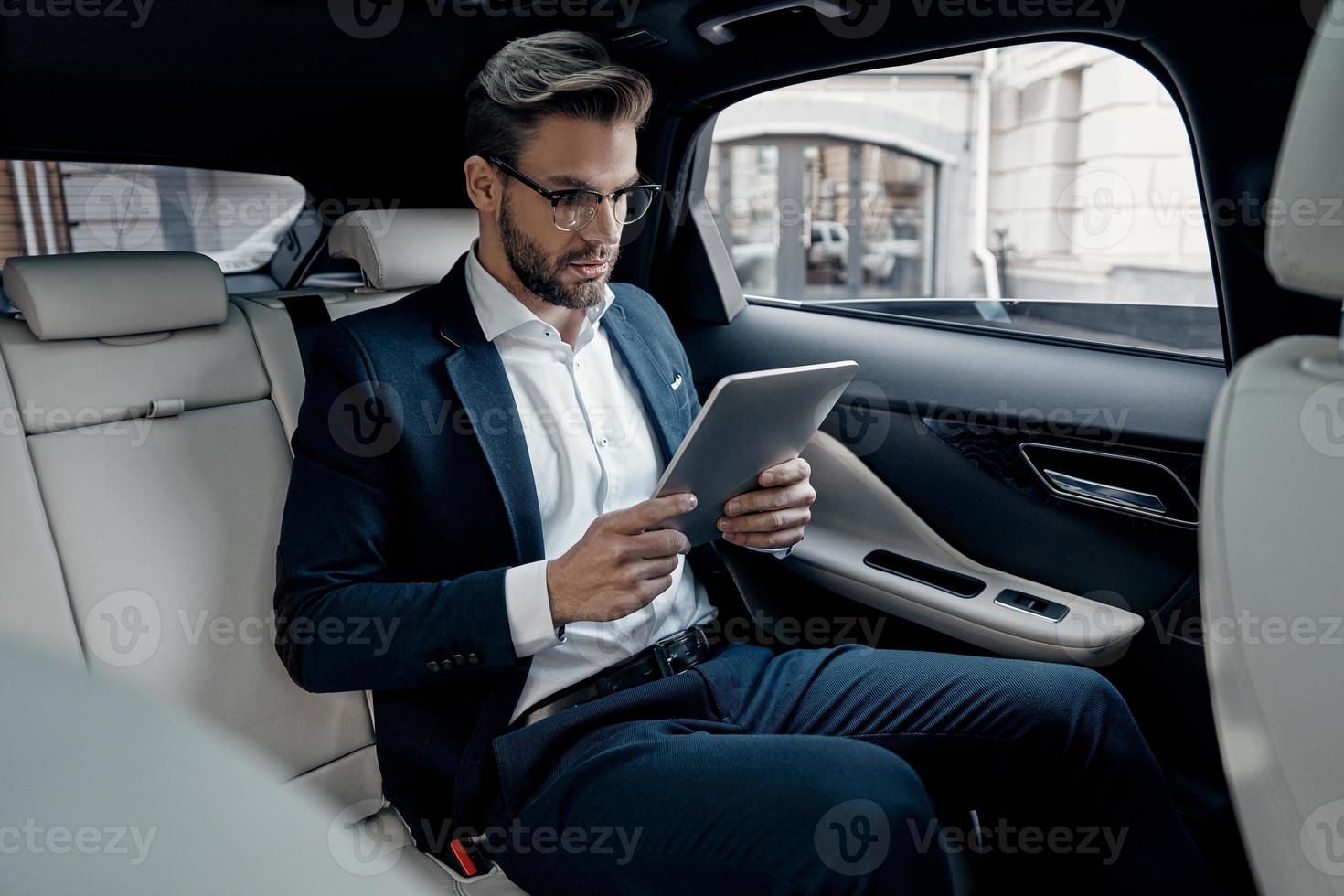 Concentrated at work. Confident young man in full suit working using digital tablet while sitting in the car photo