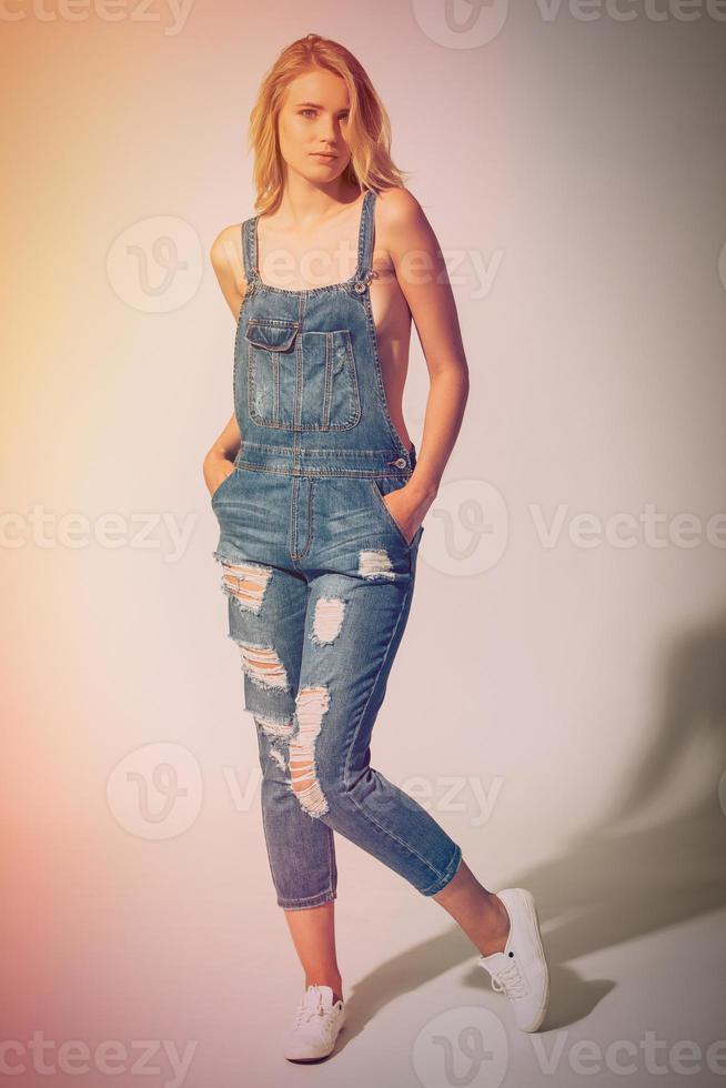 Beauty in denim style. Full length studio shot of beautiful young woman wearing jeans overall over the naked body and looking at camera photo