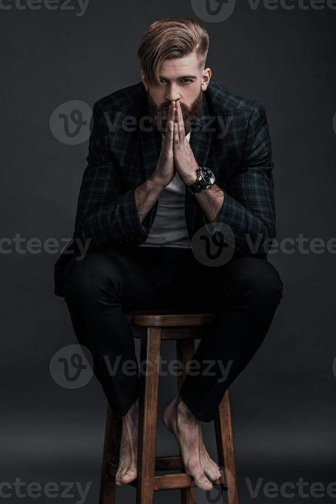 Confidence and charisma. Full length of attractive young man keeping hands clasped and looking at camera while sitting against grey background photo