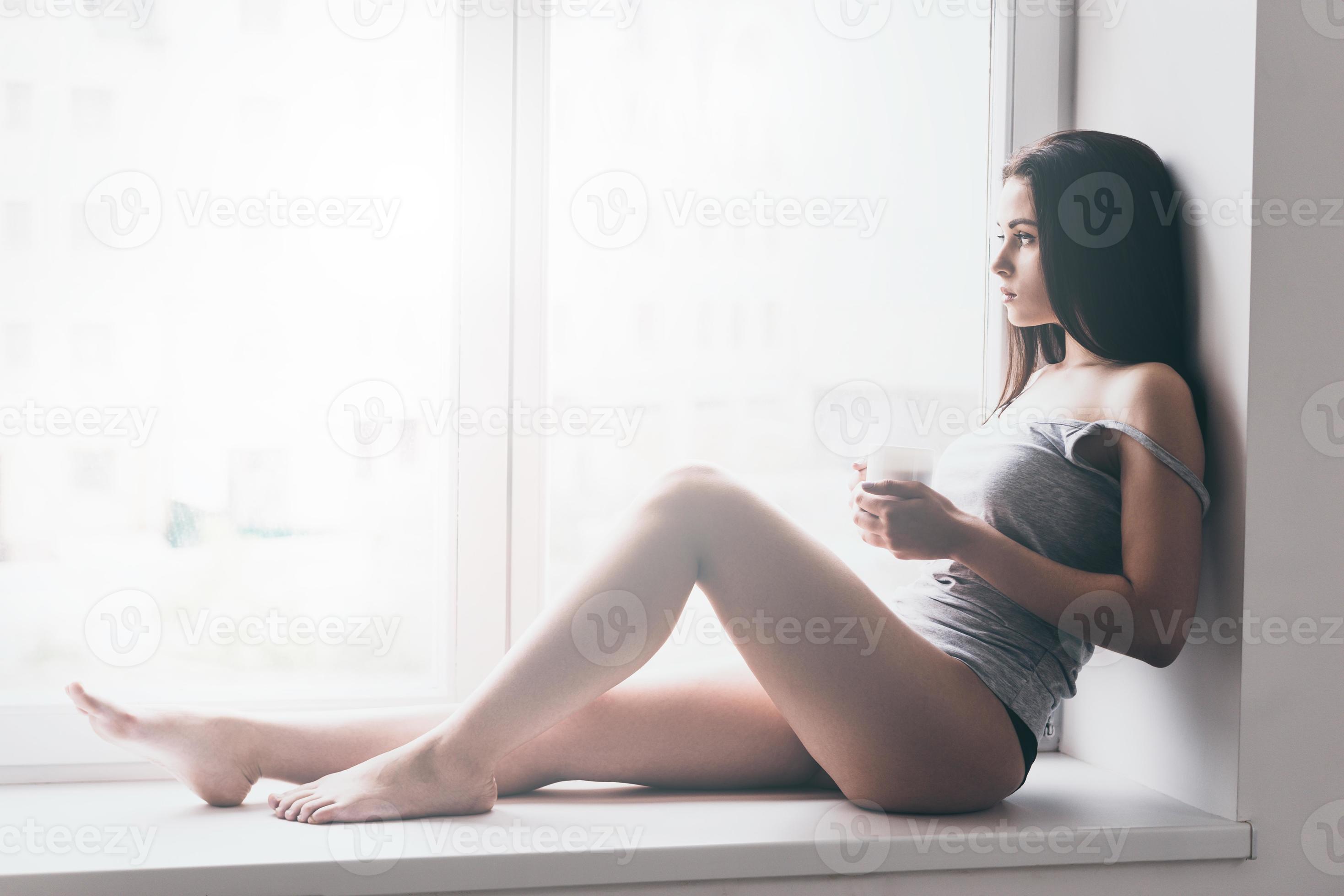 Gorgeous beauty. Beautiful young woman in panties and tank top looking  through the window while sitting on the window sill and holding coffee cup  13540508 Stock Photo at Vecteezy