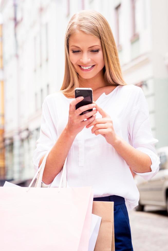 Sharing good news with friend. Close-up of beautiful young smiling woman holding shopping bags and mobile phone while standing outdoors photo