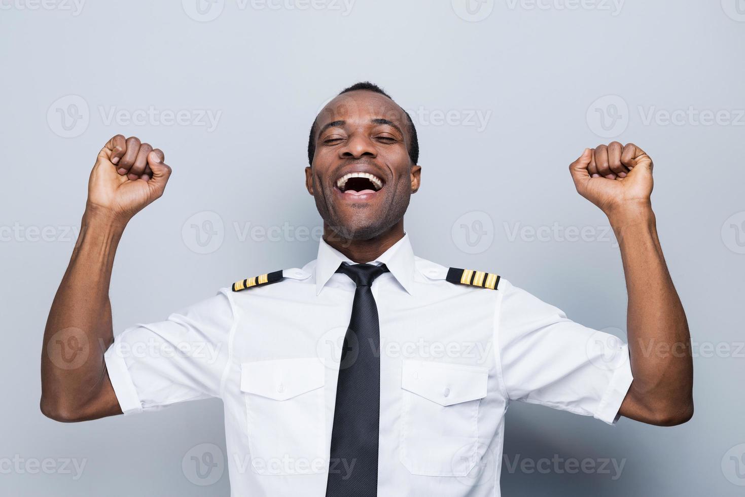 Successful pilot. Happy African pilot in uniform gesturing and keeping eyes closed while standing against grey background photo