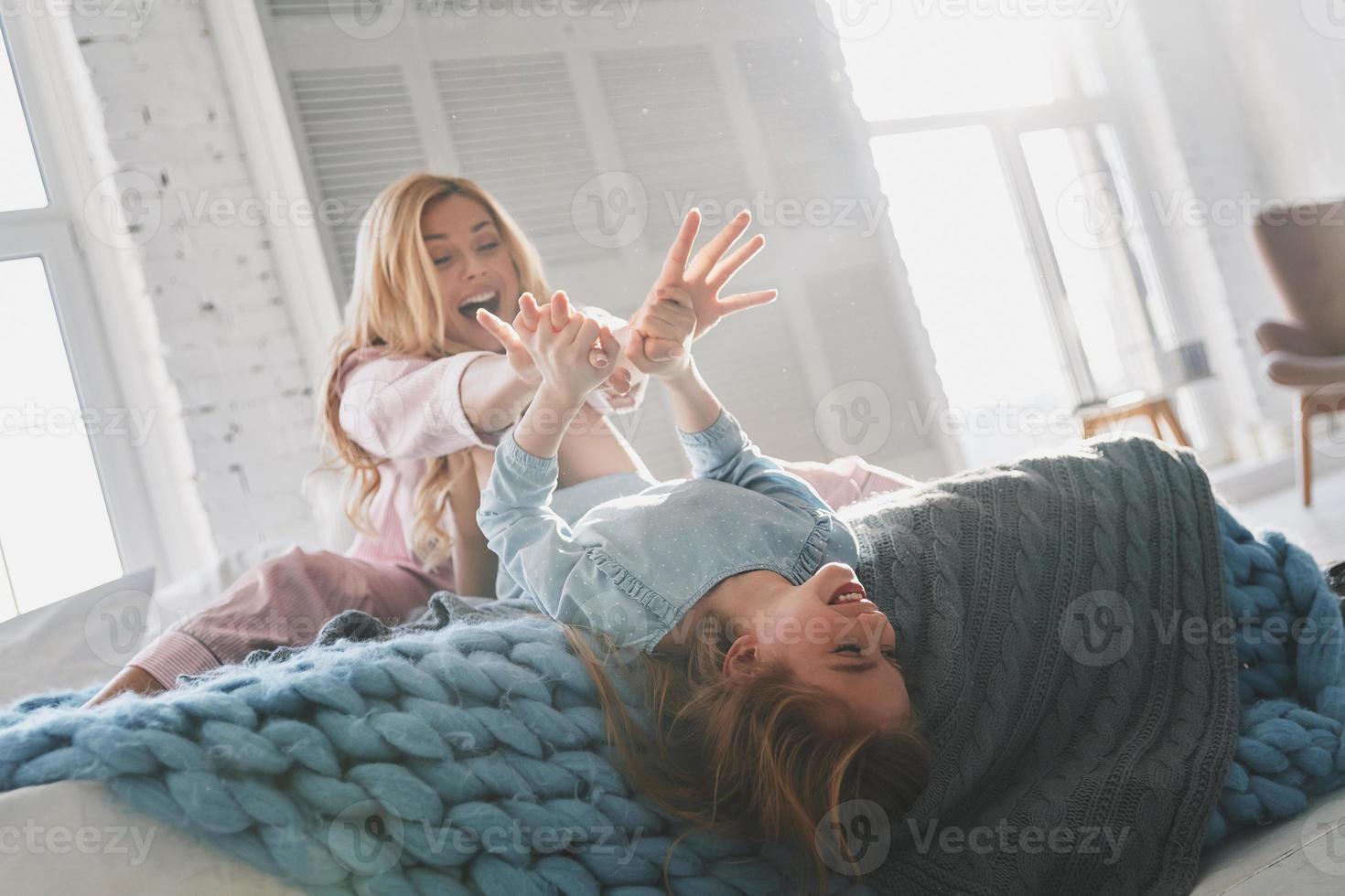 Beautiful young mother going crazy with her cute daughter and smiling while sitting on the bed at home photo
