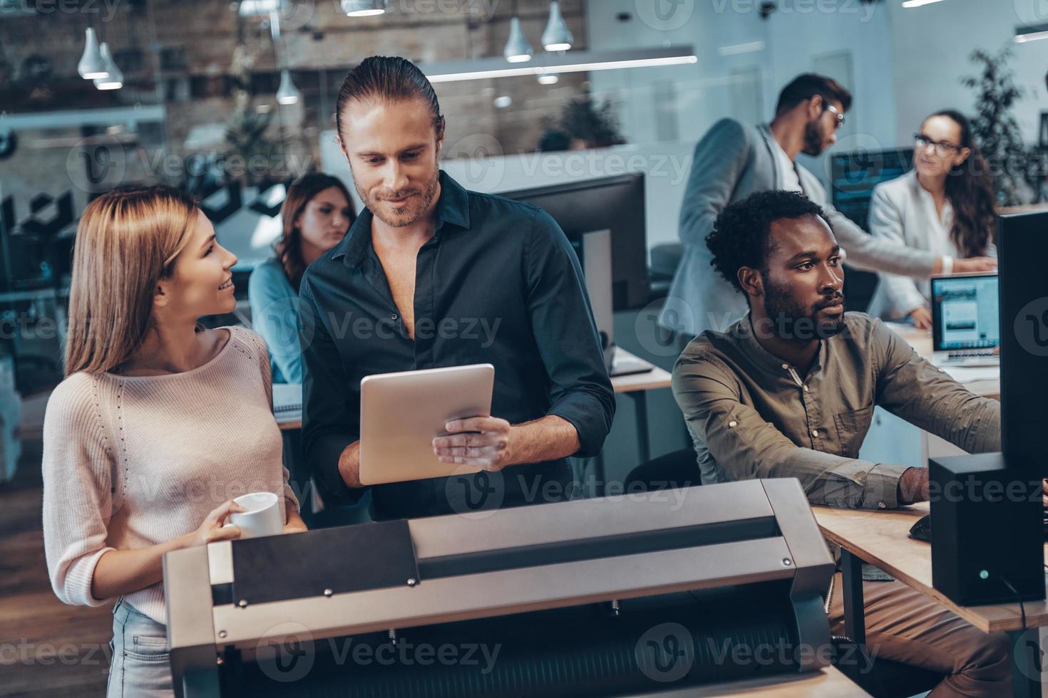 Group of young modern people in smart casual wear communicating and using modern technologies while working in the office photo